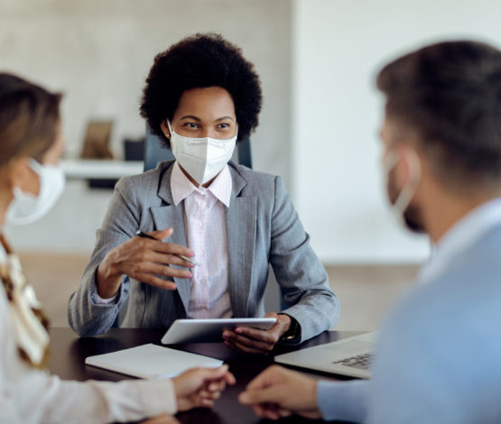 Happy black bank manager with face mask using touchpad while talking to her clients on a meeting in the office.