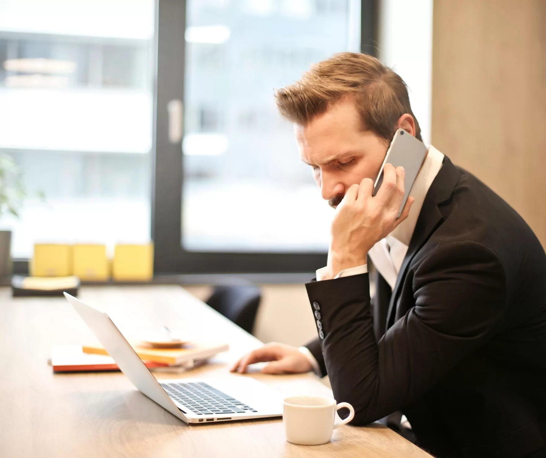 Man sitting at an office desk using a cellular phone and a laptop.